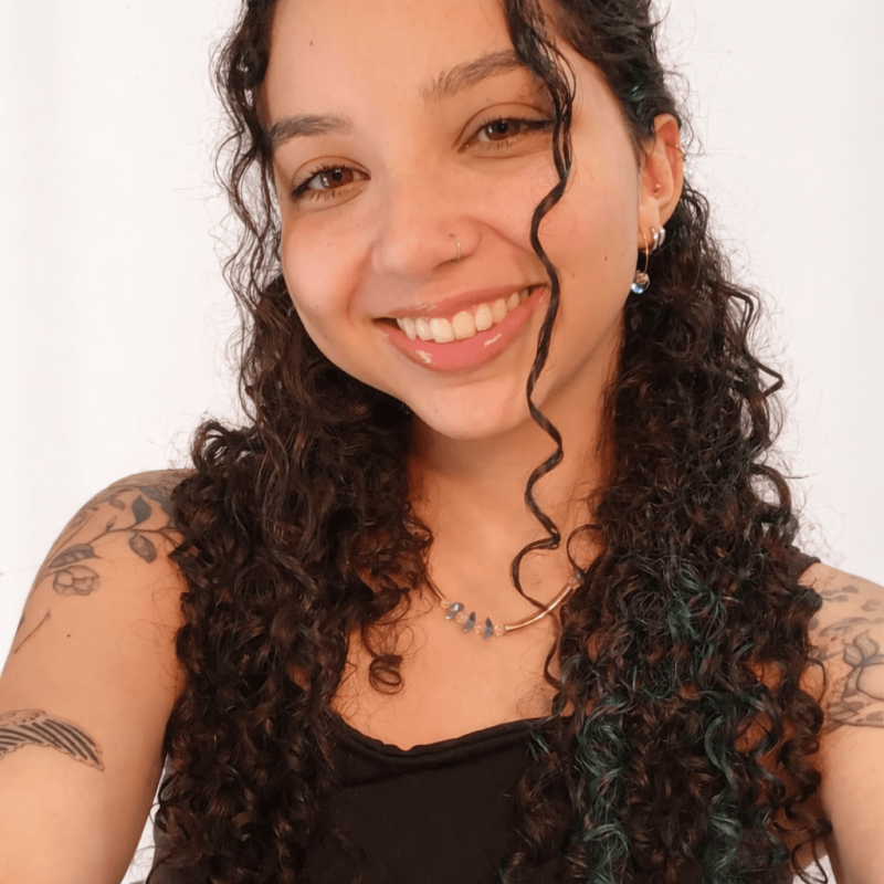 Foto de Beatriz Vitória da Silva, psicóloga com abordagem em Psicanálise na clínica Presente Psicologia. Ela está sorrindo, tem cabelos cacheados escuros e veste uma blusa branca e colar dourado.