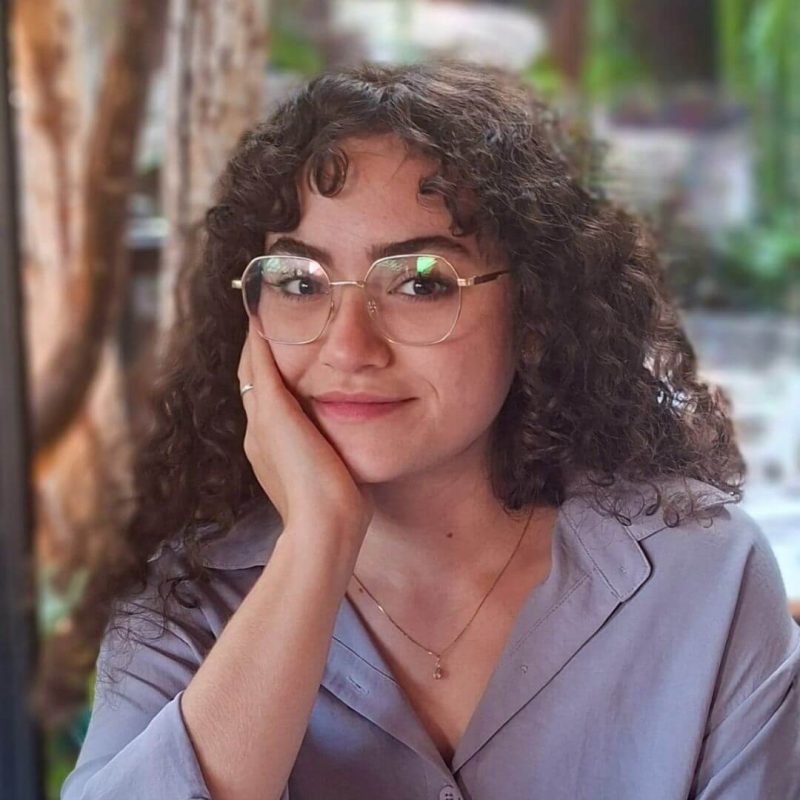 Foto da psicóloga Caroline Bezerra Oliveira, com foco em Gestalt-terapia na clínica Presente Psicologia. Ela tem cabelos cacheados castanhos, usa óculos de grau finos, veste uma camisa lilás e apoia o rosto na mão, transmitindo um sorriso leve e um olhar acolhedor.