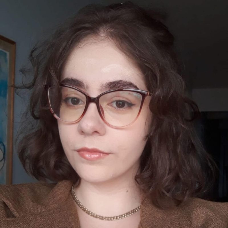 Foto da psicóloga Catarina Kuvasney Lima, especialista em Psicanálise na clínica Presente Psicologia. Ela possui cabelos castanhos ondulados na altura dos ombros, usa óculos de grau com armação escura, veste um blazer marrom sobre uma blusa branca e apresenta uma expressão serena e acolhedora.