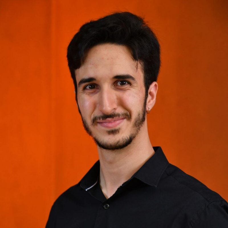 Foto do psicólogo Diogo Deganeli de Brito Varago, especialista em Análise do Comportamento na clínica Presente Psicologia. Ele tem cabelos curtos, barba aparada, sorri de forma simpática e veste uma camisa preta contrastando com um fundo laranja.