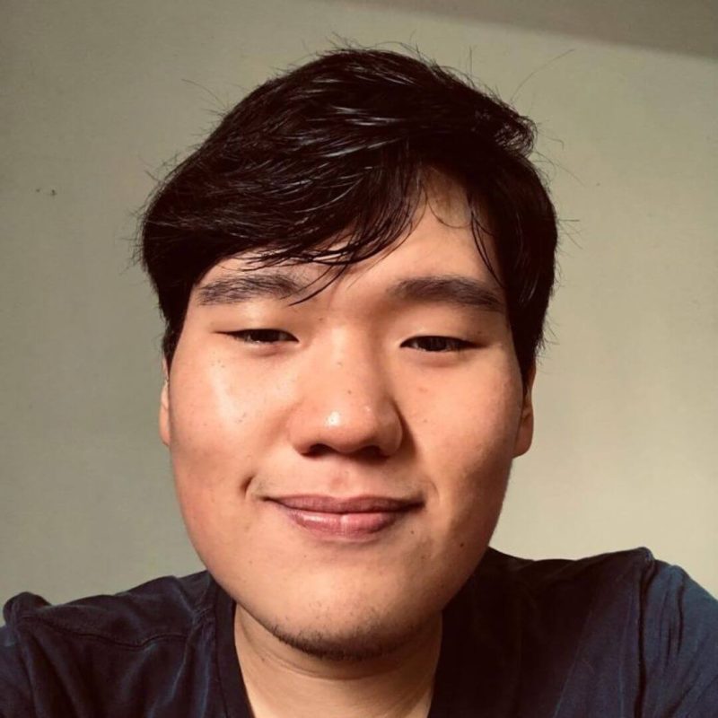 Foto do psicólogo Enrico Sumiyoshi Watanabe, especialista em Terapia Cognitivo-Comportamental e Neuropsicologia na clínica Presente Psicologia. Ele sorri de forma amigável, possui cabelos curtos escuros e veste uma camiseta azul escura.