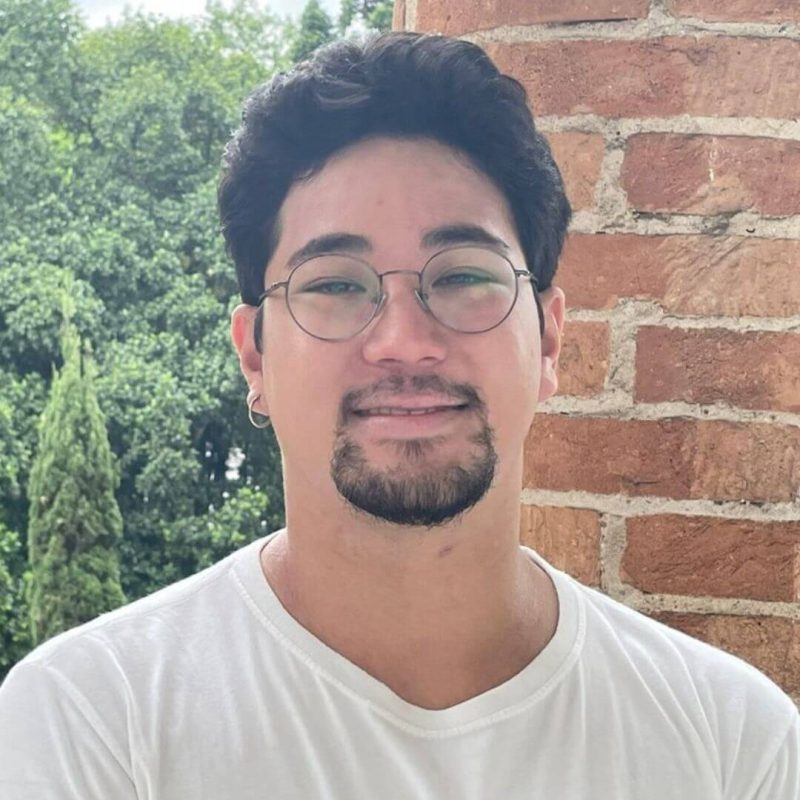 Foto do psicólogo Ian Yamamoto Mena, especialista em Terapia Cognitivo-Comportamental e Neuropsicologia na clínica Presente Psicologia. Ele veste uma camiseta branca, usa óculos de grau, possui barba e sorri de forma leve, posicionado entre duas colunas de tijolos com árvores ao fundo.