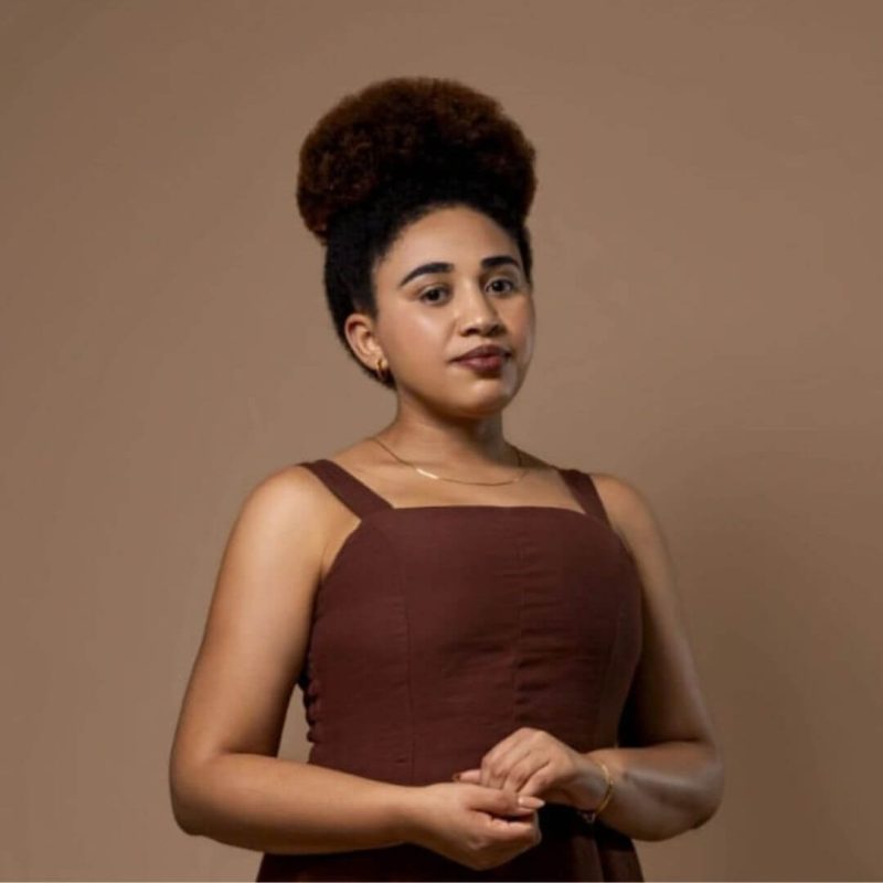 Foto da psicóloga Josefa Laura Dos Santos Paulo, especialista em Terapia Cognitivo-Comportamental (TCC) na clínica Presente Psicologia. Ela veste um vestido marrom, tem o cabelo preso em um coque elegante e posa de forma acolhedora.