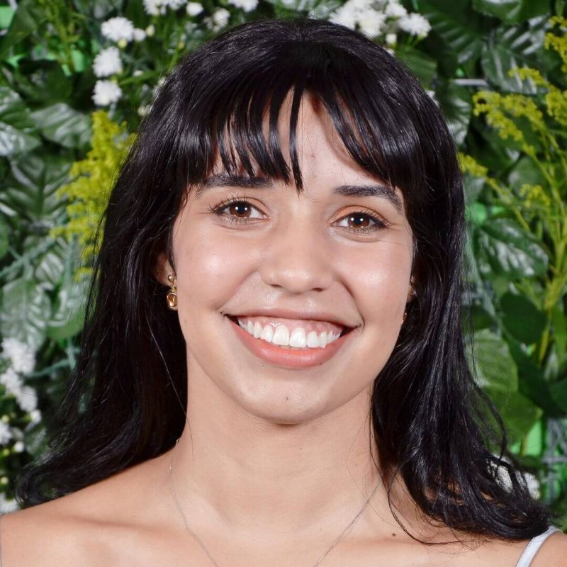 Foto da psicóloga Kamila Pereira Sousa, especialista em Psicanálise na clínica Presente Psicologia. Ela tem cabelos escuros lisos com franja, veste uma blusa clara prateada e sorri de forma amigável e acolhedora contra um fundo de folhagens verdes.
