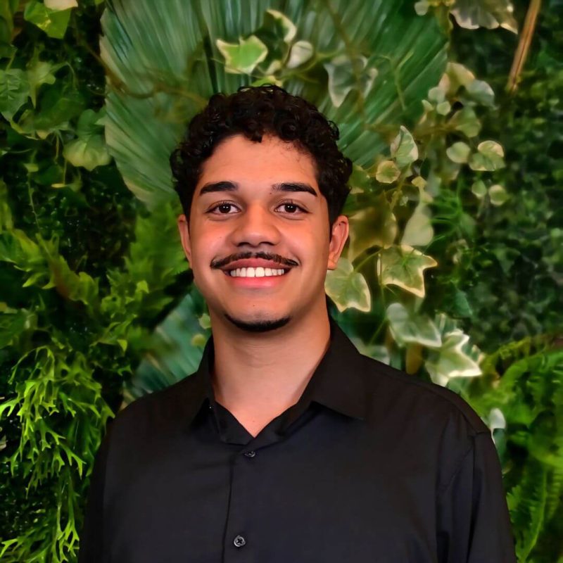Foto do psicólogo Luiz Gustavo Garcez, especialista em Terapia Cognitivo-Comportamental (TCC) na clínica Presente Psicologia. Ele sorri de forma acolhedora, veste uma camisa preta e está posicionado em frente a um fundo com folhagens verdes.