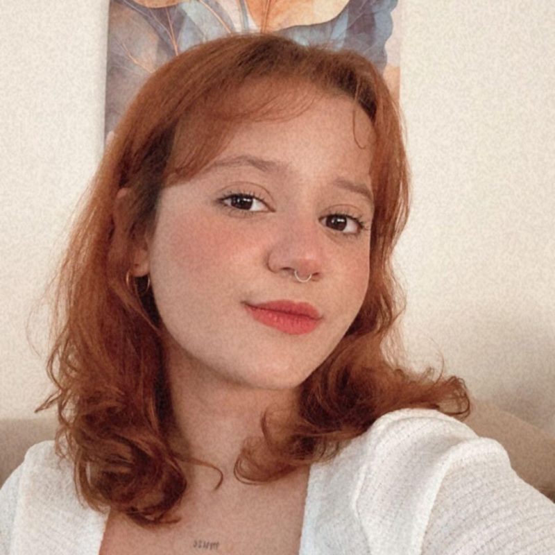 Foto da psicóloga Mayara Flores Bezerra, especialista em Análise do Comportamento e Psicopedagogia na clínica Presente Psicologia. Ela tem cabelos ruivos na altura dos ombros, usa piercing no septo, sorri levemente e veste uma blusa branca