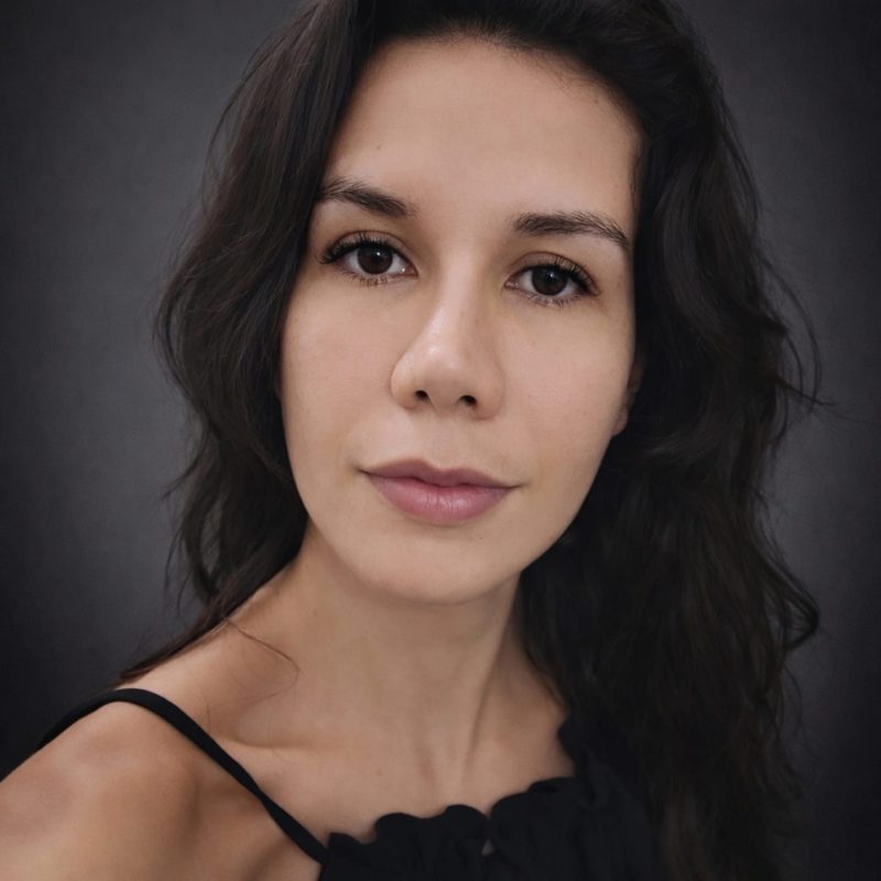 Foto da psicóloga Michelle da Silva Velasco, especialista em Gestalt-terapia na clínica Presente Psicologia. Ela tem cabelos escuros ondulados, olhar atento e sereno, vestindo blusa preta sobre um fundo escuro.