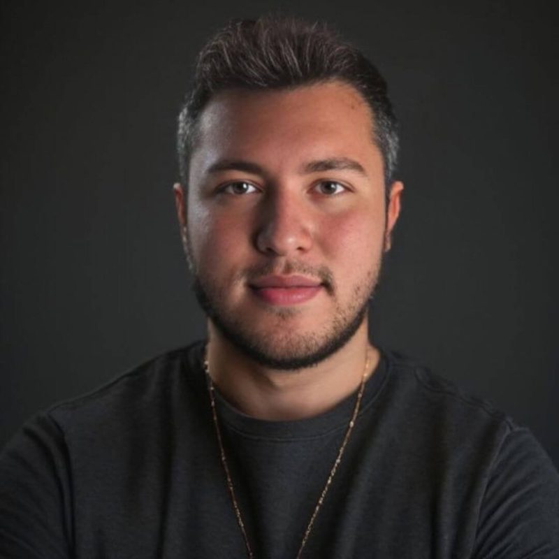 Foto do psicólogo Octávio Henriques Tavares, especializado em Psicanálise Winnicottiana e atendimento infantil na clínica Presente Psicologia. Ele sorri de forma sutil, usa barba e veste uma camiseta cinza escura com um colar dourado, posicionado em um fundo escuro.