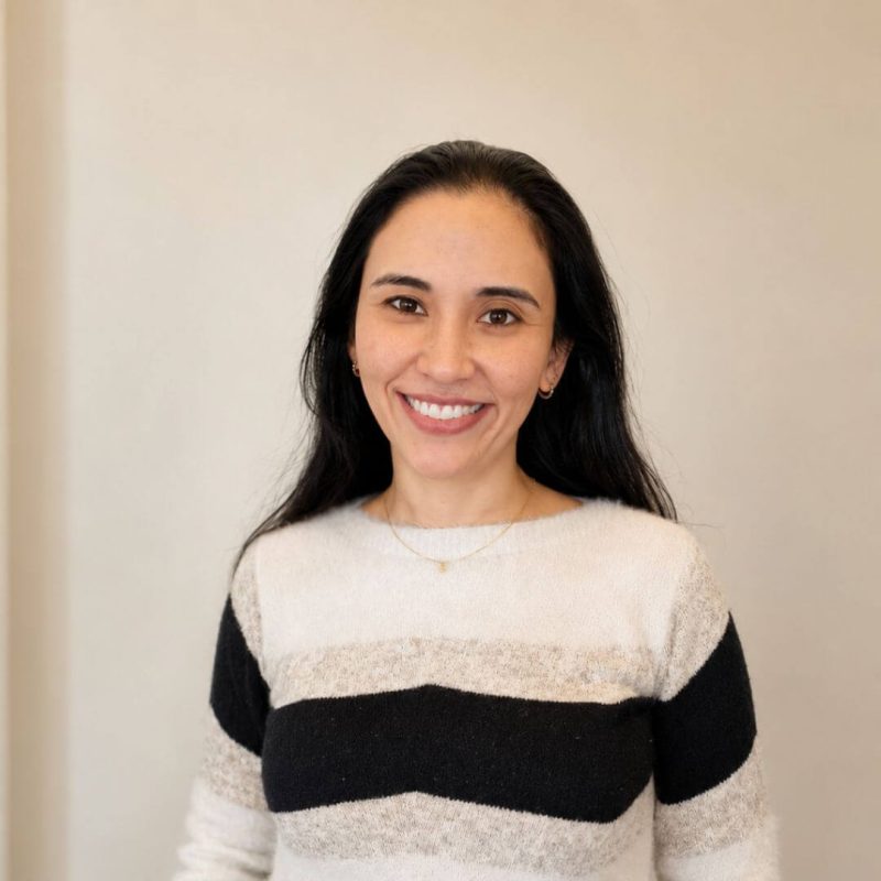 Foto da psicóloga Priscilla Tamy do Nascimento Ozono Xavier, especialista em Terapia Cognitivo-Comportamental (TCC) e Psicologia Perinatal na clínica Presente Psicologia. Ela tem longos cabelos escuros, sorri de forma acolhedora e veste um suéter com listras claras e escuras.