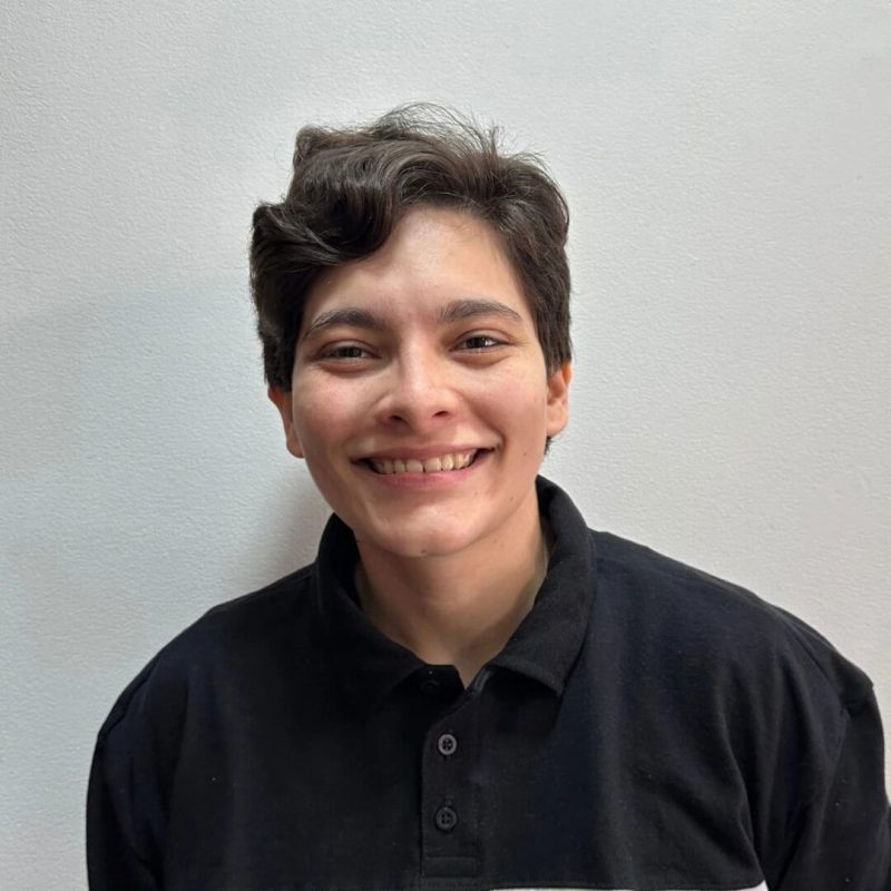 Foto da psicóloga Rebecca Scardelato Dallamarta, especialista em Psicanálise Lacaniana na clínica Presente Psicologia. Ela tem cabelos curtos e escuros, sorri de forma acolhedora e veste uma camisa polo preta contra um fundo branco.