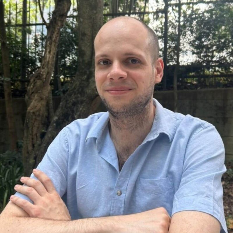 Foto do psicólogo Rodrigo Moreira Mandetta, profissional de Psicanálise na clínica Presente Psicologia. Ele sorri levemente, possui barba curta, está com os braços cruzados e veste uma camisa de botão azul-clara, posicionado em um ambiente externo com árvores e uma grade ao fundo.