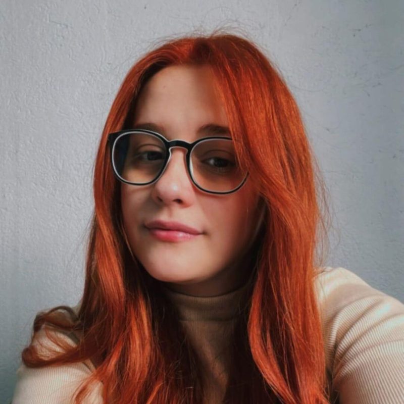 Foto da psicóloga Sabrina Kofuji Barrios, especialista em Terapia Cognitivo-Comportamental e Análise do Comportamento na clínica Presente Psicologia. Ela possui longos cabelos ruivos, usa óculos de grau com armação escura e veste uma blusa bege clara.
